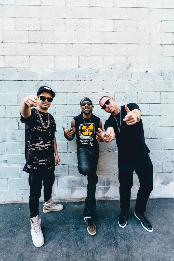 Three people pose together in front of a white brick wall