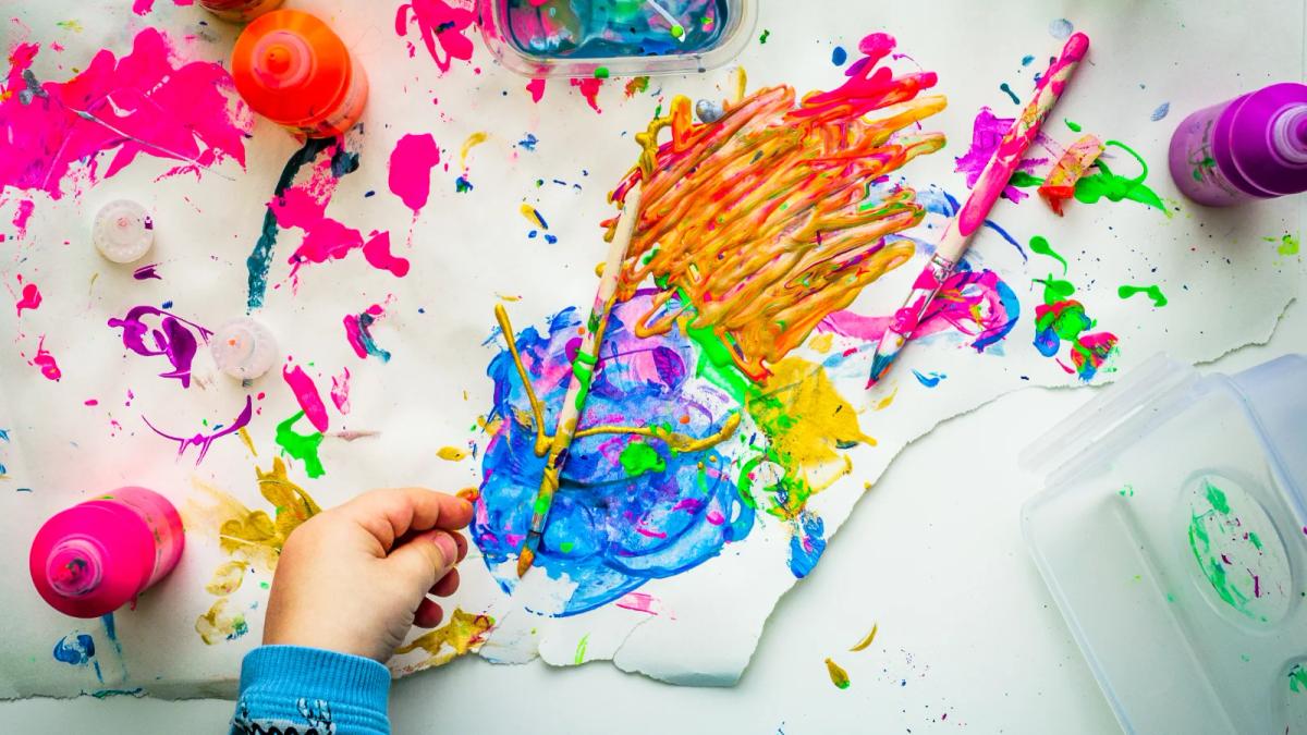 A person's hands playfully spin a colorful fidget spinner, everyday objects in the background