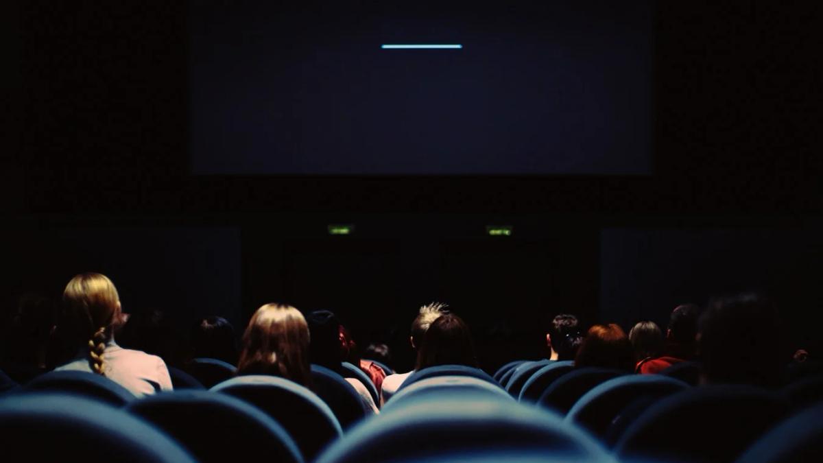 Man in theater seat with popcorn and drink, looking stressed at his phone