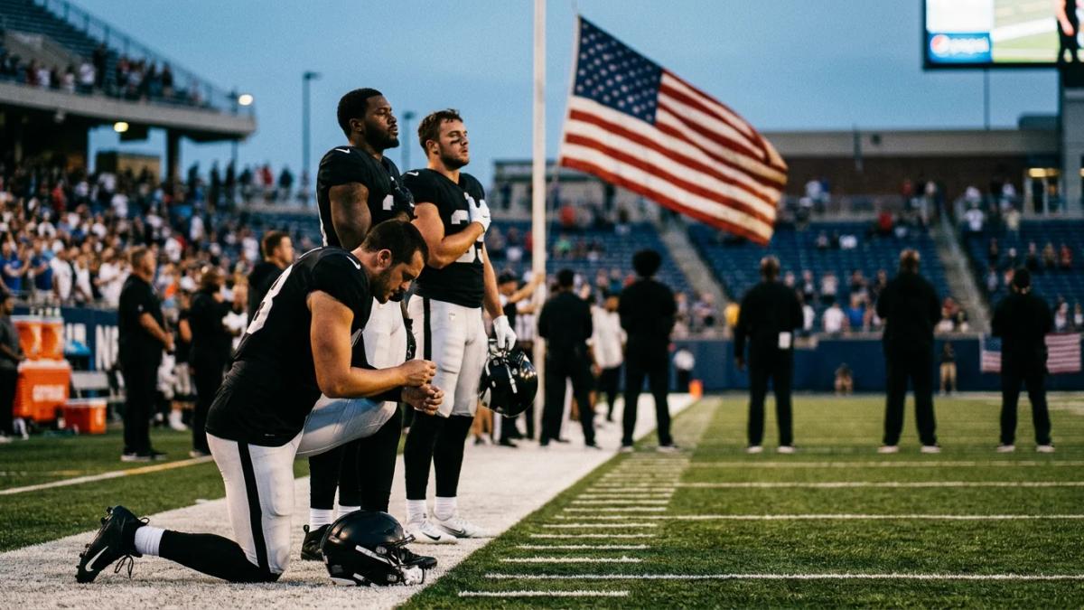 A football player kneels on the field during the national anthem with his teammates standing