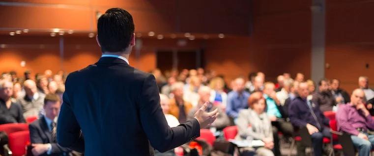 Speaker on stage looking nervous, avoiding eye contact, mic in hand, presentation slides